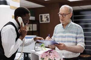African American concierge answers the telephone, caucasian retired old man signs reservation forms at hotel front desk. While receptionist is on phone call, elderly customer fills out check-in forms. photo