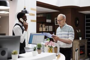 Caucasian retired old man signs reservation documents at hotel front desk as african american concierge answers call. Eldery male client filling check-in forms while receptionist speaks on telephone. photo