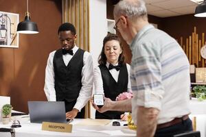 Retired senior male traveler at front desk seamlessly checking in, pays via smartphone in hotel lobby. Friendly concierge at registration counter helps old man with nfc payment using mobile device. photo