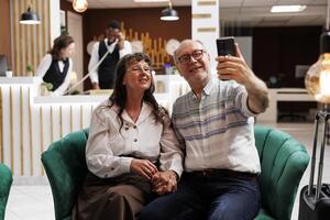 Retired senior couple videocalling family with smartphone from hotel reception. Smiling elderly male and female tourists enjoying vacation in lobby. Professional receptionists working in background. photo