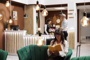 Elderly male guest arrives at front desk of hotel lobby, greeted by friendly female receptionist. Senior woman in cozy lounge area sitting on comfy sofa and reading a book while waiting for check-in. photo