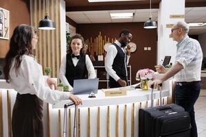 Two elderly travelers come at the hotel reception desk to check in. Multiethnic receptionists assist retired senior man and woman with booking and paperwork, therefore creating a pleasant environment. photo