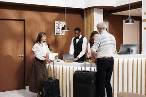 Male and female elderly guests in modern hotel lobby, with attentive staff providing efficient assistance during check-in and reservation procedures. Senior tourists wait with suitcases at front desk. photo