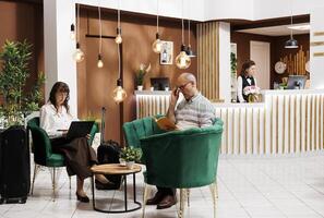 Retired elderly woman sitting on a sofa in the hotel lobby, and using her laptop. Senior male client reading a book in lounge area, while older lady is browsing on her wireless computer. photo