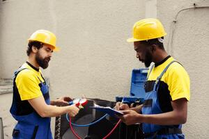Trained engineer using set of manometers to read pressure of liquids and gases in condenser unit while expert repairman writes air conditioner faulty components report list on clipboard photo