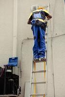 Competent mechanic in professional uniform with technical equipment climbing folding ladder to do maintenance on rooftop hvac system. Expert repairman commissioned to check up on air conditioner photo