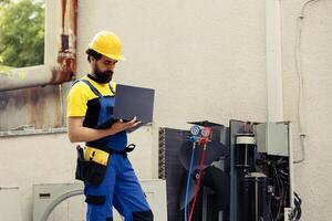 entrenado mecánico oficial para externo hvac sistema rutina mantenimiento, mirando para dañado eléctrico componentes meticuloso electricista haciendo condensador la seguridad controlar, escritura reporte en ordenador portátil foto