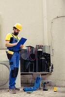 Competent wireman commissioned for outdoor hvac system routine check up, writing report on clipboard. Expert doing air conditioner inspection and safety check, looking for faulty components photo