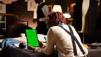 Female tailor working with greenscreen on tablet, using isolated chroma key display in textile workshop. Woman holding device with blank copyspace on screen, handmade tailoring. photo