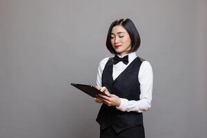 Restaurant administrator young asian woman using digital tablet while posing in studio. Attractive waitress wearing professional uniform and bow tie holding portable device photo