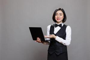Smiling asian waitress in professional uniform showing laptop and looking at camera. Young attractive receptionist pointing with hand while holding portable computer portrait photo