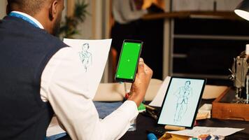 Tailor using greenscreen in atelier with team of designers, looking at isolated display on smartphone. Male couturier working with blank chroma key template on screen, tailoring concept. photo