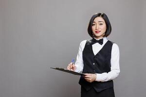 Young asian waitress in uniform holding clipboard, writing customer order and looking at camera. Attractive woman receptionist holding pen and checklist studio portrait on gray background photo