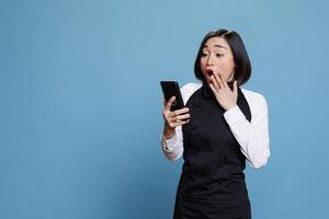 Excited asian woman receptionist covering mouth with surprised gesture while reading client positive feedback on smartphone. Cafe shocked waitress receiving good news on mobile phone photo