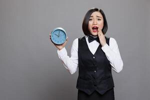 Anxious asian waitress panicking while holding circle dial alarm clock and looking at camera with open mouth. Young terrified woman receptionist showing timer watch portrait photo