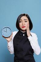Upset asian waitress showing alarm clock and looking at camera with disappointed facial expression. Sad woman receptionist checking time while running late studio portrait on blue background photo