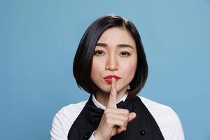 Hotel young asian woman receptionist making silent gesture, asking to keep secret portrait. Restaurant waitress wearing uniform holding forefinger on lips and looking at camera photo