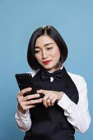 Young smiling asian waitress wearing restaurant uniform tapping on smartphone touchscreen. Attractive woman receptionist answering client message while using mobile phone photo