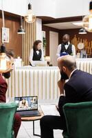 Entrepreneurs using laptop for online call, meeting clients in hotel reception lobby while they are travelling on work trip. Businessmen talking to people on remote videoconference chat. photo