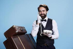 Visitors calls hotel telephone and discuss with refined bellboy about bookings, studio background. Skilled hotel receptionist uses corded phone to talk about commodities and concierge services. photo