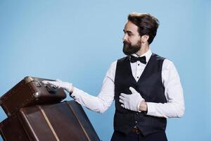 Male bellhop points left and right to create new advertisement, indicating direction sideways while he wears formal attire. Young classy doorman presents ad over blue background. photo