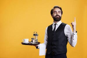 Waiter with serving tray points upwards on camera, showing something up above head while he wears professional restaurant clothes. Young man butler suggesting direction in studio. photo