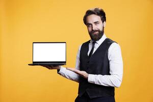 Professional man holds laptop with white display in studio, presenting isolated copyspace template on camera. Young adult employee using pc with blank empty mockup screen, yellow background. photo