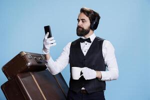 Modern bellhop checks messages and holding cup of coffee in studio, enjoying break with music on headset. Young man with professional hotel industry job texting and drinking cold brew. photo