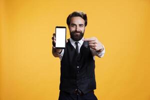 Confident waiter points to white screen on camera, presenting isolated mockup blank display on mobile phone. Young butler with suit and tie shows empty device with copyspace template. photo