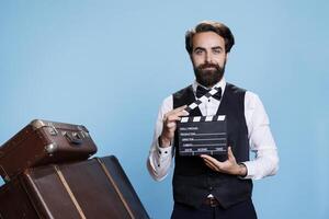 Doorkeeper clapping filmslate on camera, saying action to cut scene for fun against blue background. Classy hotel bellboy pretending to work in movie production using clapperboard. photo