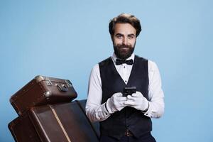 Hotel porter scrolls on smartphone, using social media and texting messages while he poses in studio with trolley bags. Doorman bellhop in suit browsing online page on phone app. photo