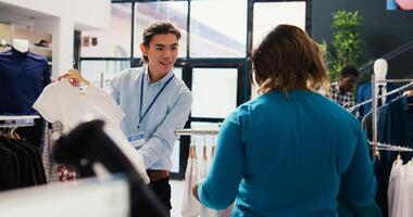Asian worker helping customer to buy trendy clohtes, discussing shirt fabric in modern boutique. African american woman shopping for fashionable merchandise, looking at new fashion collection photo