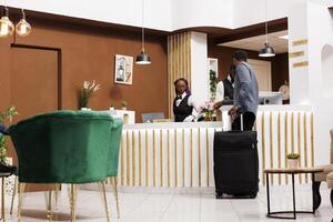 Hotel check in process. African American man traveler with suitcase standing at reception desk talking with receptionist registering his arrival, tourist waiting for key or access card to room photo