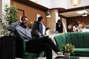 Happy smiling young African American man traveler sitting in hotel lobby having call while waiting for check-in. Tourist holding digital tablet, waving hand greeting friend looking at camera photo