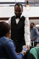 Cashless hotel. African American woman guest making contactless payment using credit card while resting in lobby. Smiling waiter holding pos terminal accepting payment over nfc technology photo