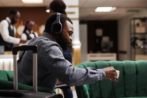 Serious African American businessman wearing headphones drinking coffee while waiting for hotel room to be ready, sitting with luggage in lobby. People and traveling, business trip concept photo