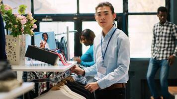 Asian worker arranging new fashion collection, putting hangers with fashionable merchandise on racks in modern boutique. Employee preparing store for opening, working with stylish clothes photo
