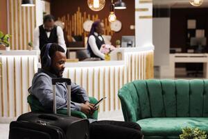Smiling young African American man traveler waiting for room in hotel. Black guy in headphones using digital tablet while sitting in lobby with luggage, watching movie, wait for check-in process photo