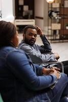 Young African American guy wheelchair user traveling with companion, disabled man tourist sitting in hotel lobby talking with girlfriend or wife while waiting for check-in, traveler with disabilities photo