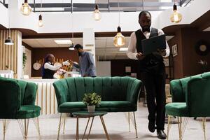 Young African American man hotel employee wearing standing uniform with laptop in lobby, using computer software program to make reservations. CRM in the hospitality industry photo