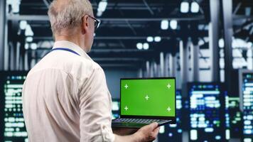 Seasoned IT professional navigating network of server cabinets in industrial hub. Expert with chroma key laptop ensuring proper operations, doing optimizations and maintenance in data center photo