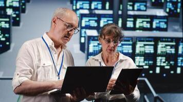 Knowledgeable IT practitioners looking around server farm, using laptop and tablet to crosscheck disaster recovery plan and assess rackmounts in need of replacement, preventing perils photo