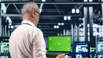 Capable IT professional navigating network of server clusters in industrial hub. Expert with green screen laptop ensuring proper operations, doing optimizations and maintenance in data center photo