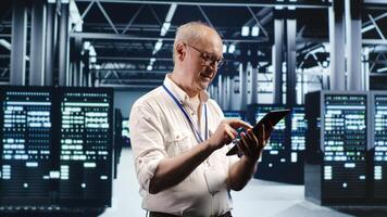 Older professional navigating network of server rigs in industrial cloud computing business. Worker with tablet doing scans and optimizations in high tech data center, preventing system worries photo