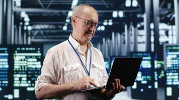 Certified IT professional navigating network of servers in industrial space. Admin with laptop ensuring flawless operations, doing optimizations and maintenance in data center photo