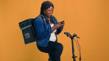 Female courier relaxing with smartphone on bicycle on break from delivery service. Enthusiastic african american woman checking cell phone while waiting for online restuarant order for delivering. photo