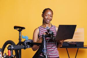 Sporty african american lady repairing broken bicycle with assistance from internet on digital laptop. Youthful black woman examining and fixing damaged components with toolkit and personal computer. photo