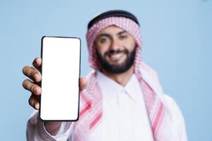 Man in arabic attire holding smartphone displaying white screen for application advertising. Smiling muslim person posing in studio and showing mobile phone blank touchscreen closeup photo