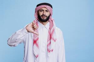 Muslim man dressed in traditional arabian clothes showing thumb down studio portrait. Arab in thobe and ghutra showcasing disagreement and looking at camera with serious expression photo