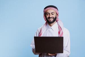Smiling man wearing traditional muslim clothes standing with laptop and browsing internet. Cheerful arab using portable computer and typing online message while posing in studio photo